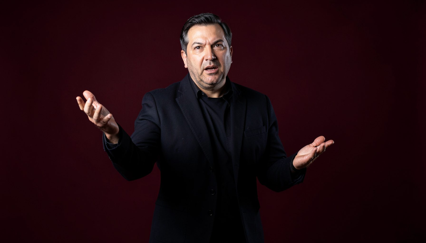 A conductor in a dark jacket gesturing expressively against a deep burgundy backdrop