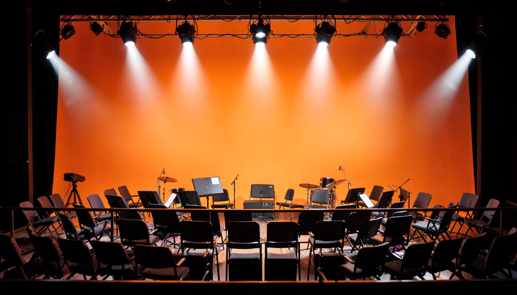 A concert hall stage seen from the balcony, with orchestra chairs arranged under bright white spotlights