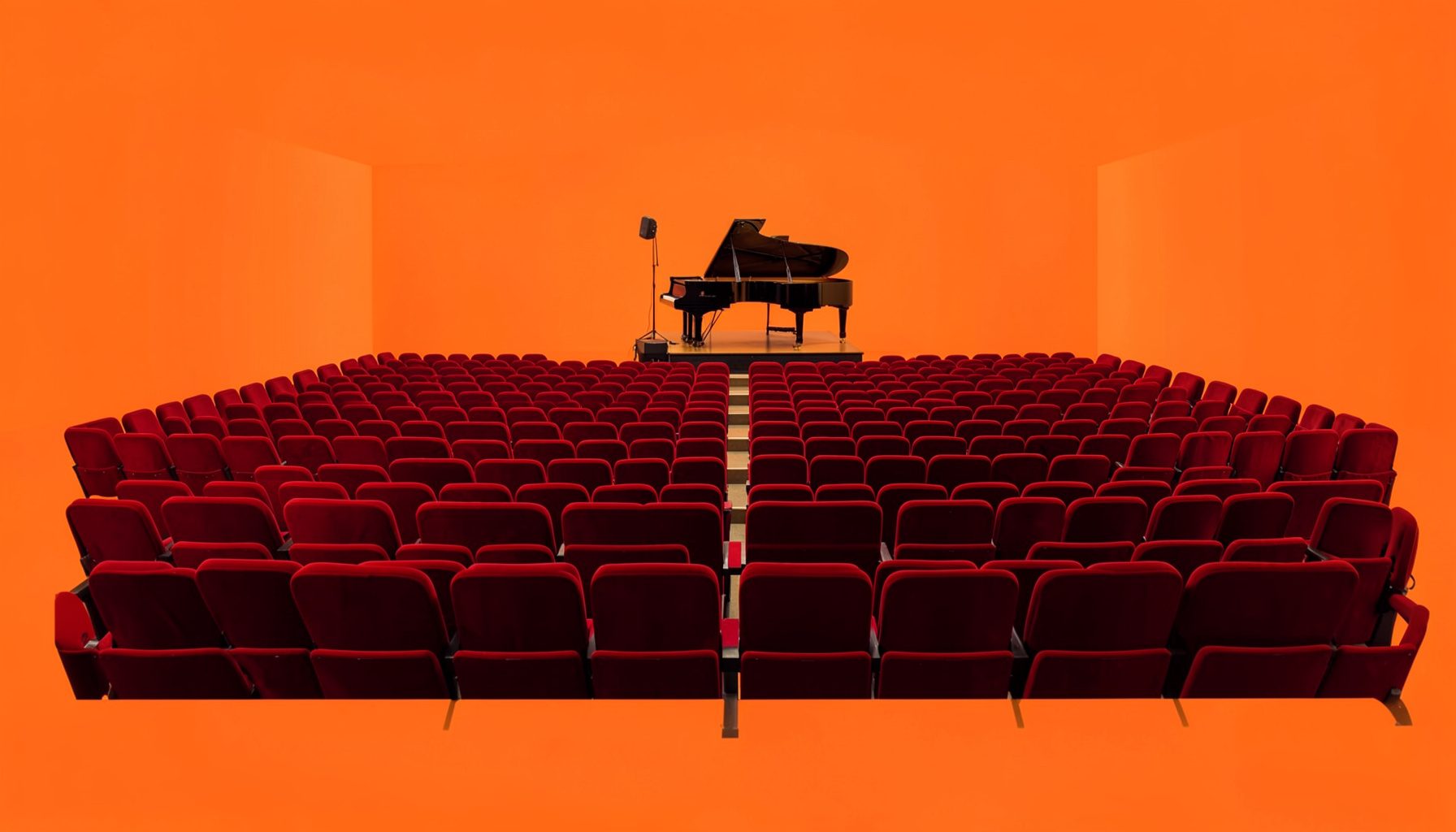 A flat document-style photograph of a concert hall interior with rows of empty red velvet seats and a grand piano on stage